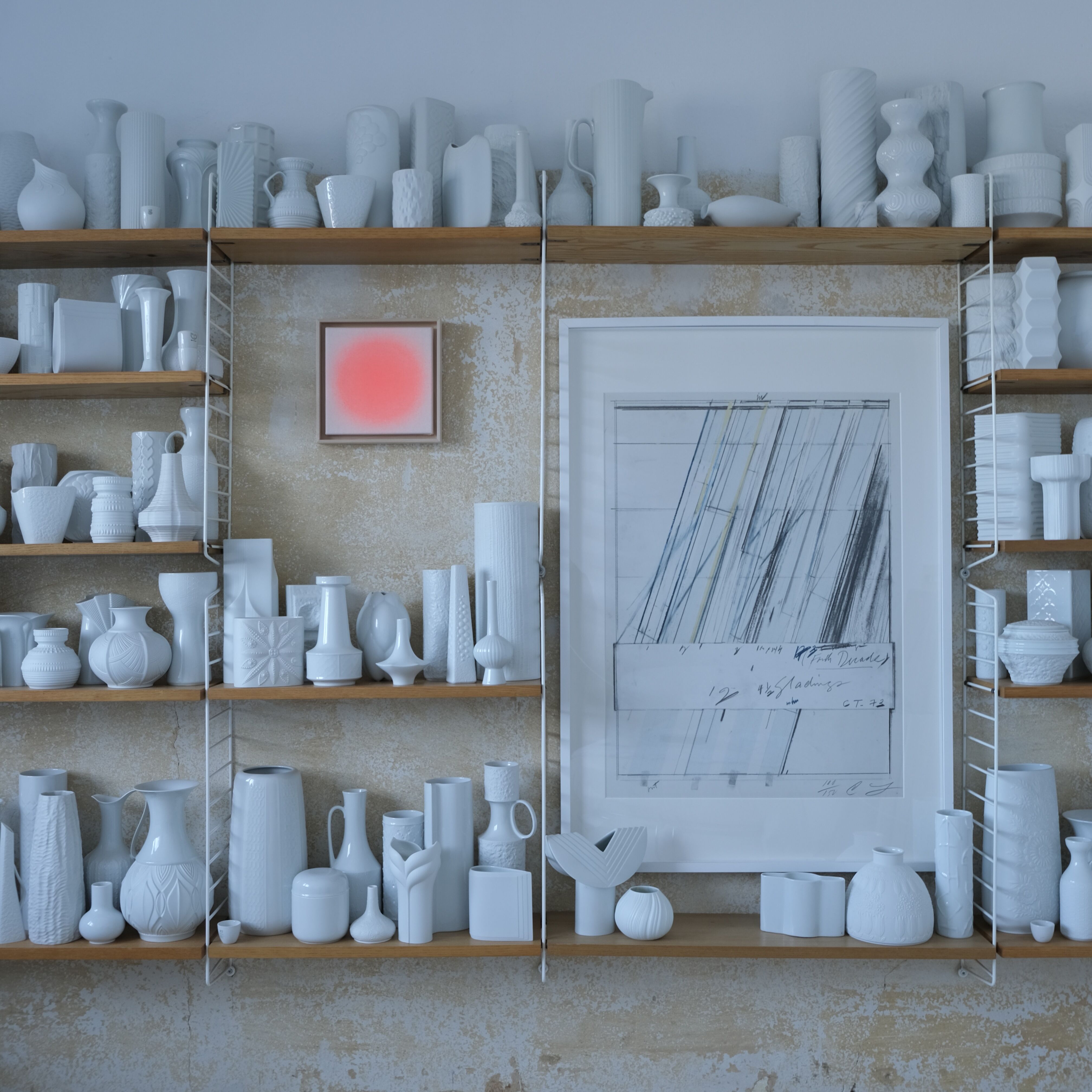 Detailreiche Ansicht eines offenen Wandregals mit vielen weißen Keramikvasen und Dekoobjekten auf hölzernen Böden vor einer beigen Wand. Zwischen den Regalböden hängt die gerahmte ZEIT-Sonderedition Sorbet »Apricot« von Anastasia Benko – ein zart orangefarbenes, quadratisches Acryl-Pigmentkunstwerk.