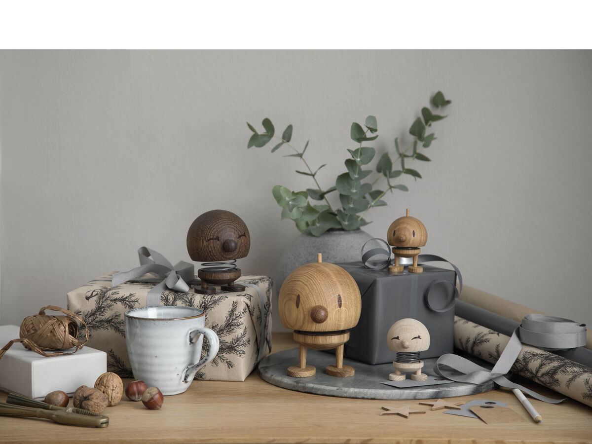 Vier Hoptimist Bumble Figuren aus Holz auf einem Tisch. Zwei Geschenkpakete, ein grauer Blumentopf mit Eukalyptus, eine weiße Tasse und Walnüsse sind ebenfalls zu sehen. Hintergrund ist eine graue Wand.