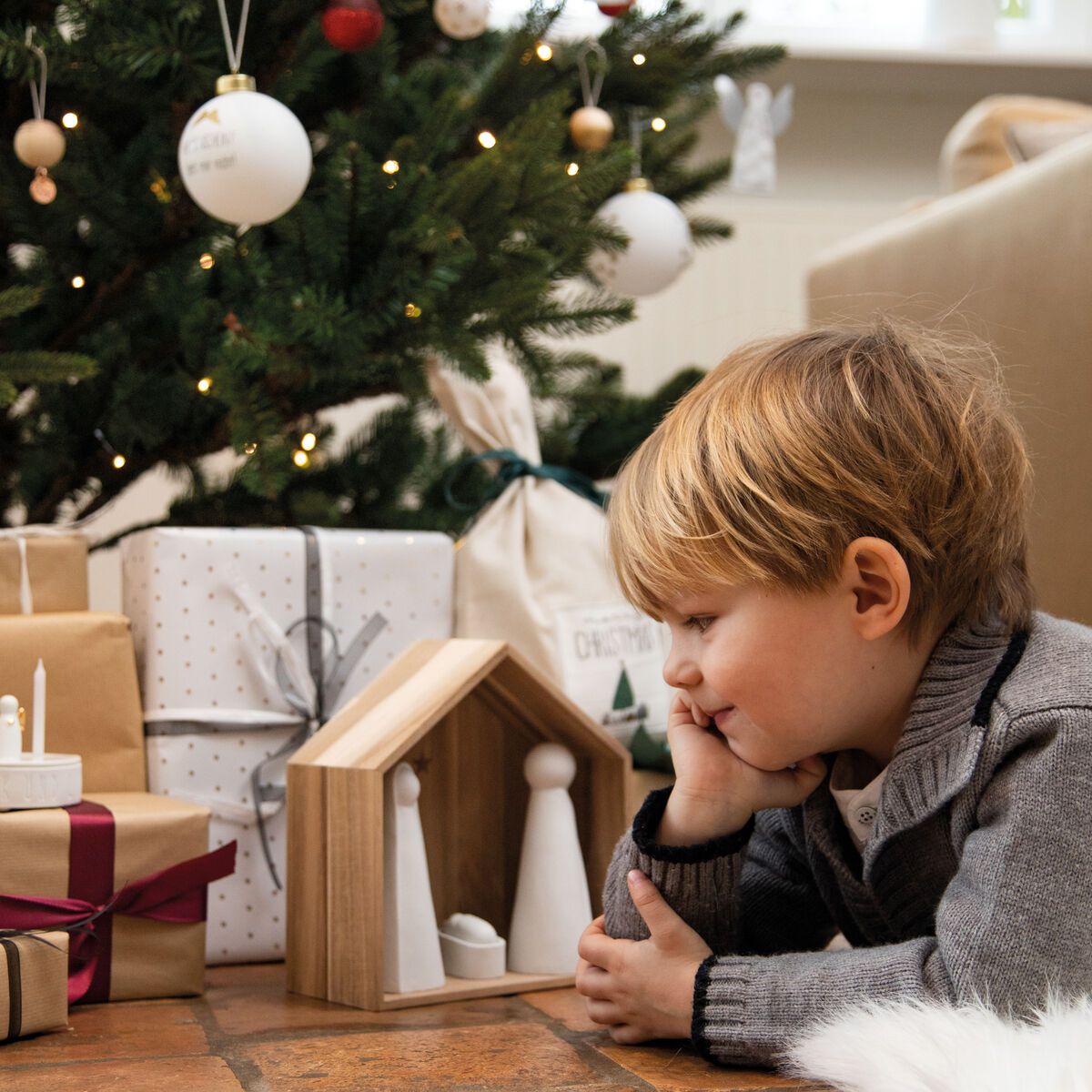 Ein Kind liegt auf dem Boden vor einer Holz-Weihnachtskrippe mit minimalistischen Figuren. Im Hintergrund steht ein geschmückter Weihnachtsbaum mit Lichtern und Kugeln, daneben liegen eingepackte Geschenke.