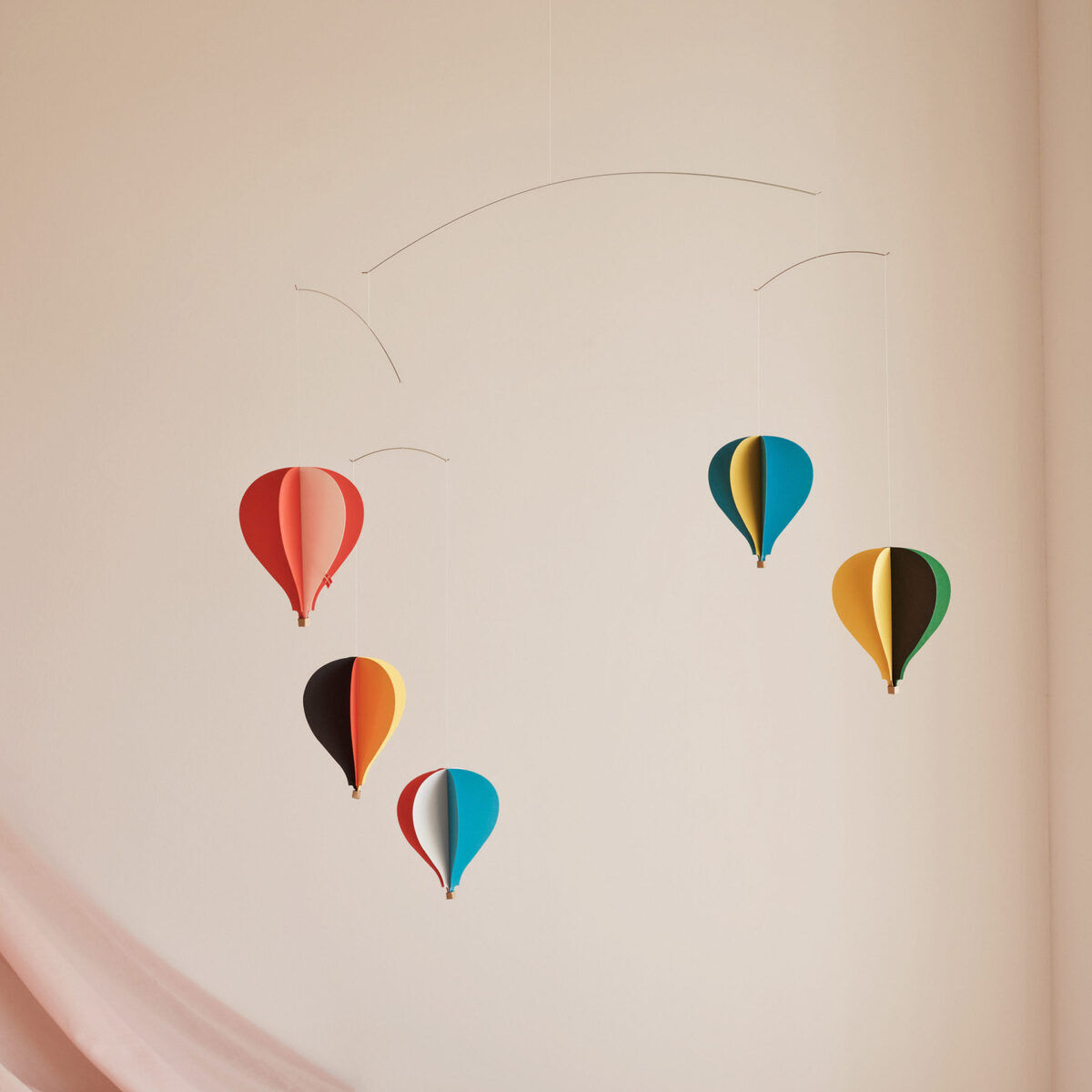 Ein Mobile mit fünf bunten Heißluftballons hängt an einer Wand. Die Ballons sind jeweils in Rot, Blau, Gelb und Grün gehalten. Im Hintergrund ist ein Teil eines rosafarbenen Tuchs zu sehen, das von einer Ecke des Bildes hängt.