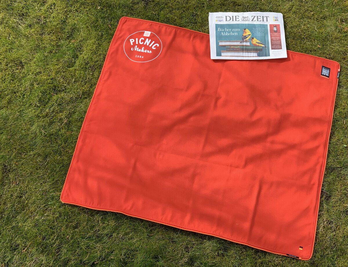 Eine rote Picknickdecke mit dem Aufdruck »Picnic «Squarepark 11« liegt auf grünem Gras. Auf der Decke liegt eine Zeitung.