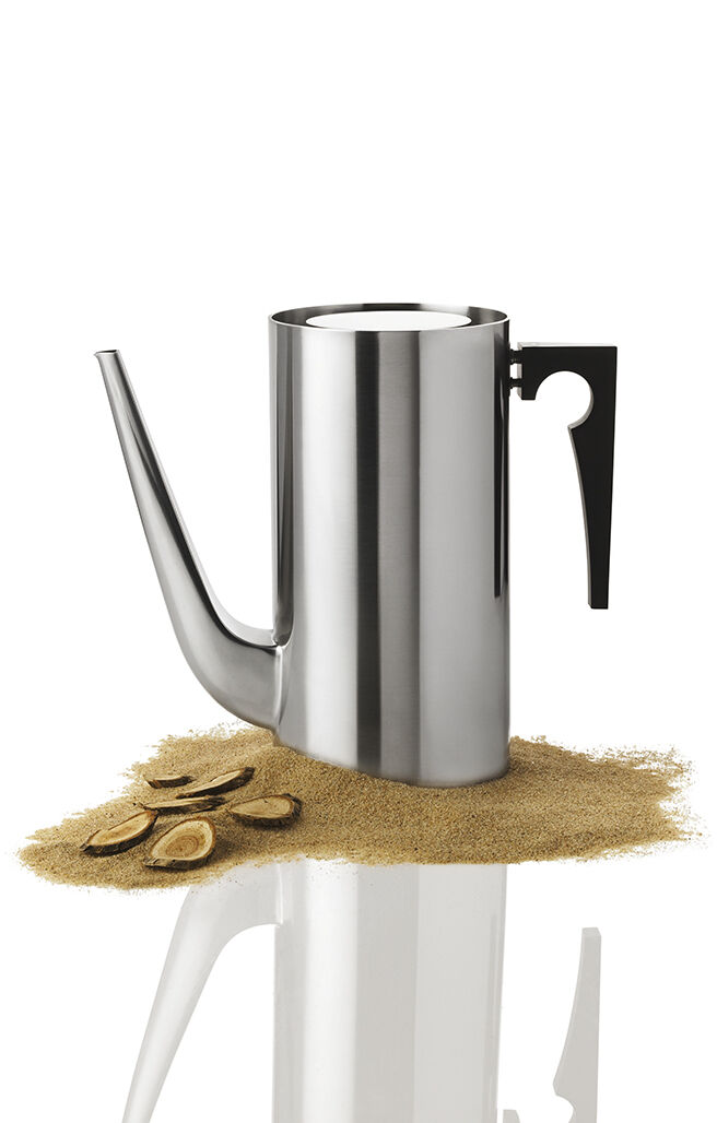 Eine elegante Kaffeekanne aus Edelstahl, entworfen von Arne Jacobsen, auf einem weißen Hintergrund. Die Kanne hat ein minimalistisches und funktionales Design mit einem schwarzen Griff und einer langen Ausgusstülle. Sie steht auf einer Oberfläche mit verstreutem Kaffeepulver und Kaffeebohnen.