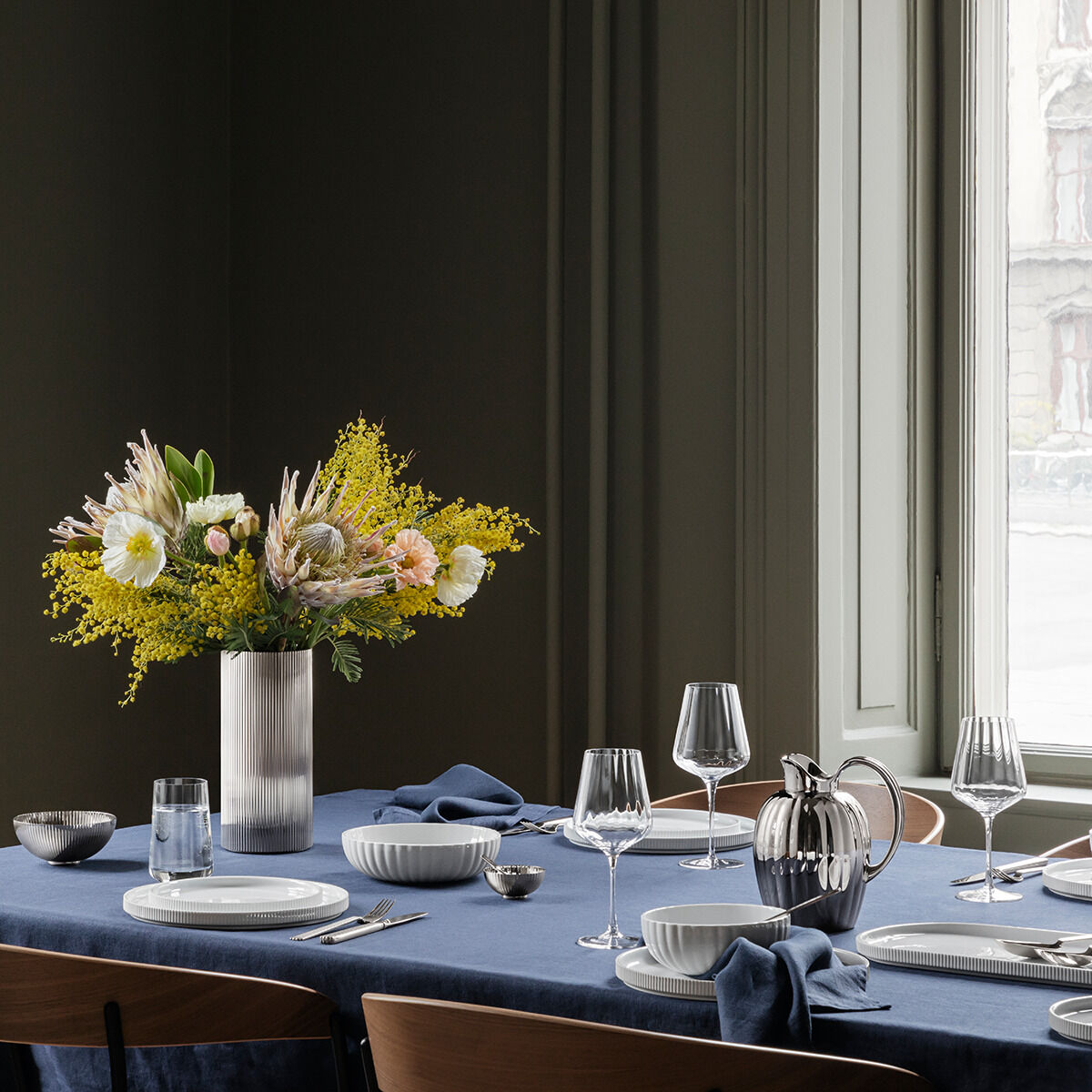 Eine elegante Vase namens »Bernadotte« steht auf einem gedeckten Tisch. Die Vase enthält ein stilvolles Blumenarrangement mit gelben und weißen Blüten. Der Tisch ist mit blauen Tischdecken, weißen Tellern, Gläsern und Besteck dekoriert. Im Hintergrund ist ein Fenster und weitere Dekorationen sichtbar, die eine moderne und einladende Atmosphäre schaffen.