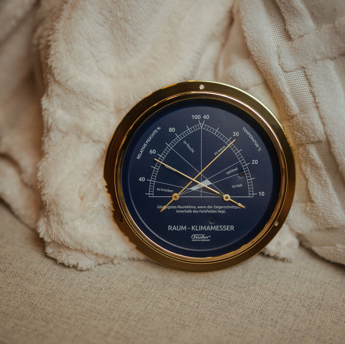 Rundes Raumklimamesser der ZEIT Sonderedition mit poliertem Messinggehäuse und blauem Zifferblatt, lehnt an einem cremefarbenen Kissen und einer weichen Decke auf einem hellen Sofa. Die Skala zeigt Temperatur- und Luftfeuchtigkeitswerte an.