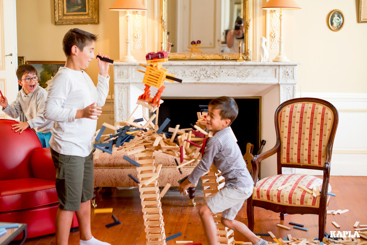 Kinder spielen mit Holzbausteinen, bauen Strukturen in einem Wohnzimmer mit Kaminsims und einem Sessel im Hintergrund. Verschiedene Holzteile sind am Boden verteilt, während die Kinder in freudiger Interaktion sind.