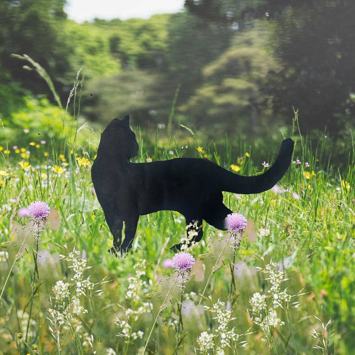 Schwarzer Gartenstecker in Form einer stehenden Katze in einer grünen Wiese mit violetten Blumen und umgeben von natürlicher Vegetation.