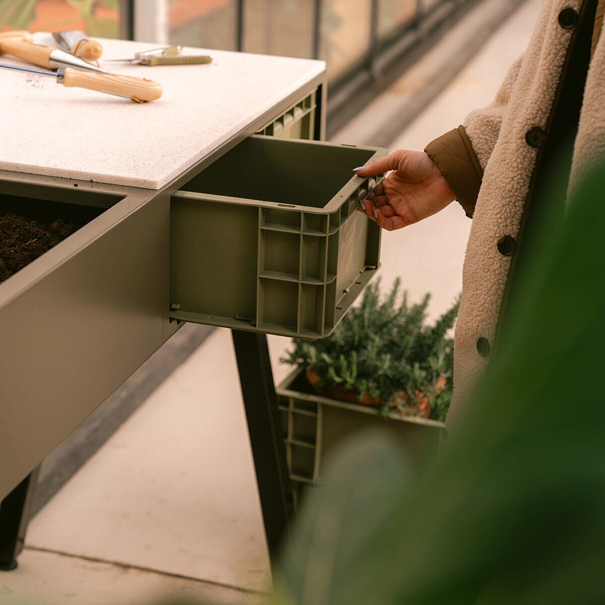Detailansicht des grünen Pflanztischs »Gardenworks Station« von Weltevree mit ausziehbarer Schublade, Werkzeugen und Pflanze im Hintergrund, genutzt von einer Person im beigen Mantel in einem modernen Gewächshaus.