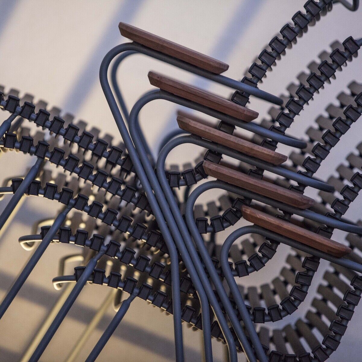Ein Stapel von CLICK Dining Chairs mit Armlehnen in einem modernen und minimalistischen Design. Die Stühle haben einen Metallrahmen und Holzelemente an den Lehnen, die Farben Schwarz, Braun und Grau sind präsent.