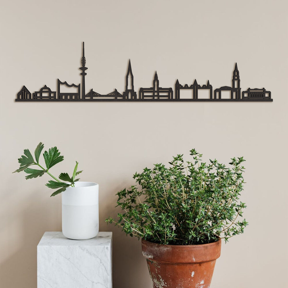 Schwarze Holzkontur der Skyline von Hamburg an einer beige Wand, mit einem weißen Ziergegenstand auf einem kleinen Sockel links und einer Topfpflanze rechts.