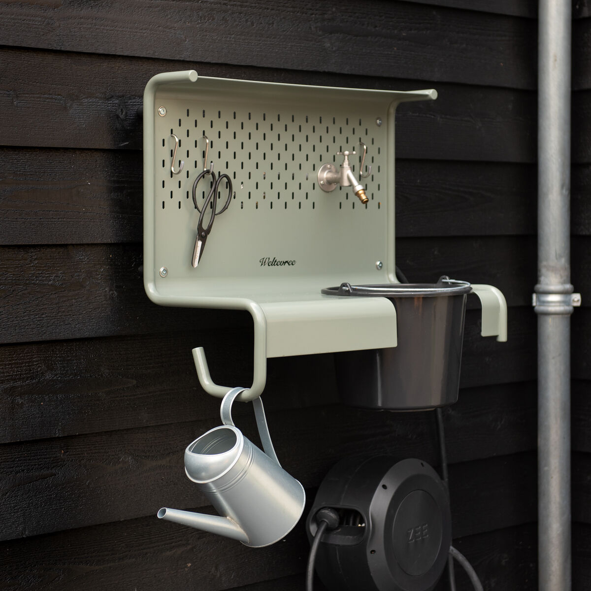 Die Gartenstation »Waterworks« von Weltevree ist an einer dunklen Holzaußenwand montiert. Auf der hellgrauen Metallkonstruktion hängen Gartenschere und Handschuhe, darunter befinden sich eine Gießkanne und ein schwarzer Eimer. Seitlich ist ein Wasserschlauch angeschlossen.