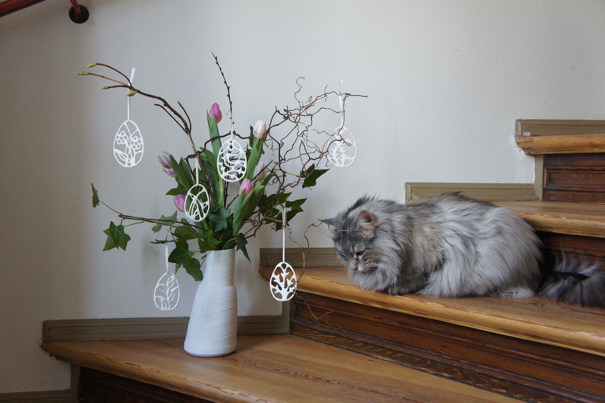 Ein Arrangement bestehend aus einer weißen Vase mit blühenden rosa Tulpen und dekorativen Zweigen, an denen weiße Porzellan-Ostereier als Anhänger aufgehängt sind. Eine graue Katze sitzt neben der Vase auf einer Holztreppe in einer Wohnung.