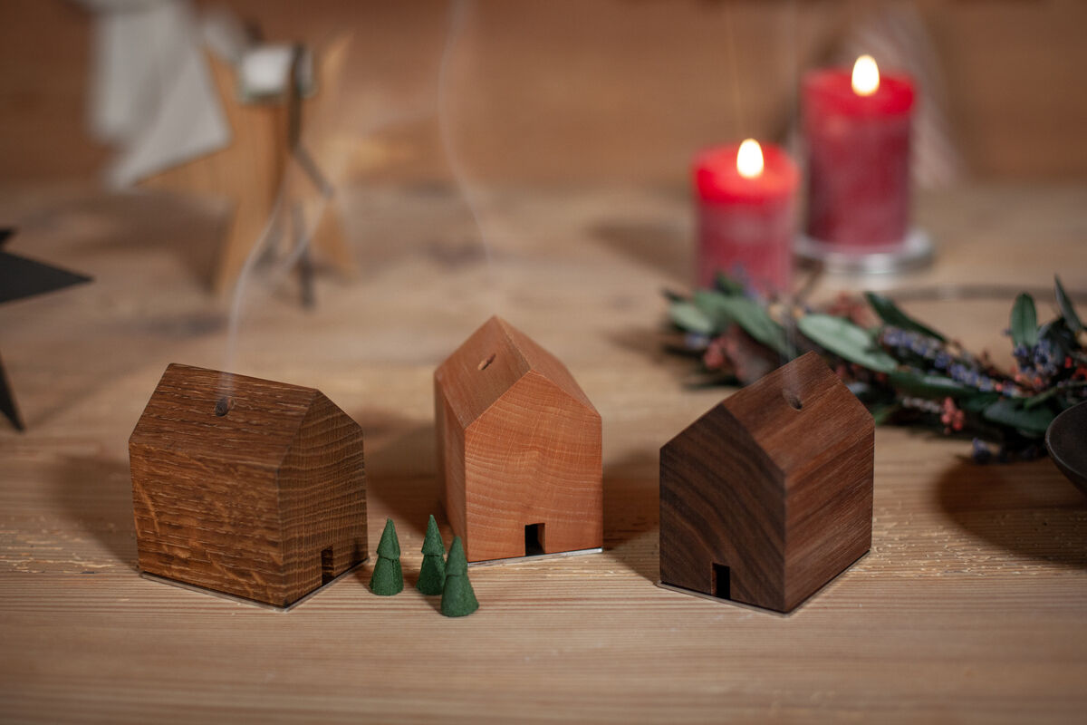 Drei kleine hölzerne Räucherhäuser, aus denen Rauch aufsteigt, umgeben von grünen Räucherkerzen und roten brennenden Kerzen, auf einem Holztisch arrangiert.