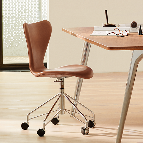 Moderner Drehstuhl mit braunem Sitz und verchromtem Gestell neben einem Holztisch mit Büchern und Brille.