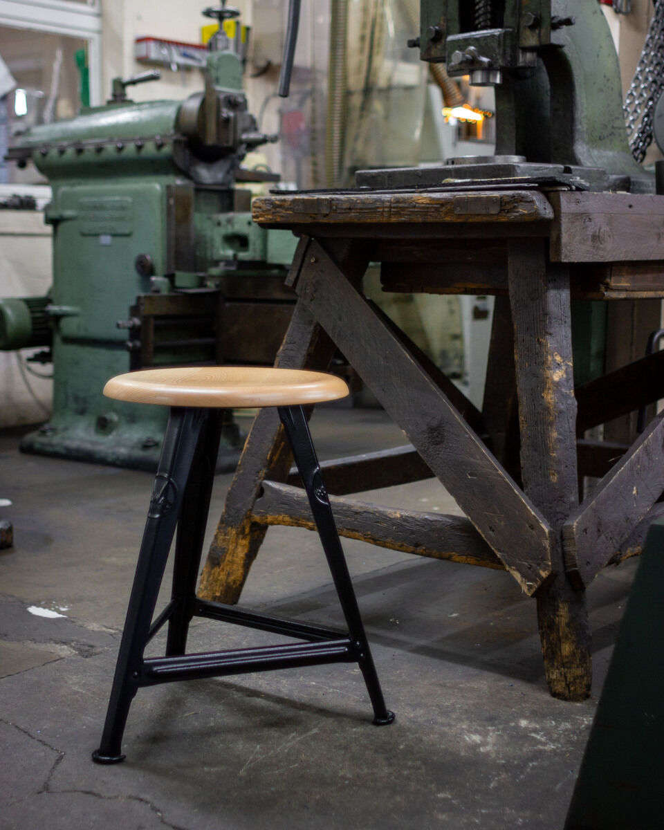 Ein industrieller Hocker mit einer eichenfarbenen Sitzfläche und einem mattschwarzen Metallgestell steht in einer rustikalen Werkstatt neben einem alten Holztisch und großen Maschinen.