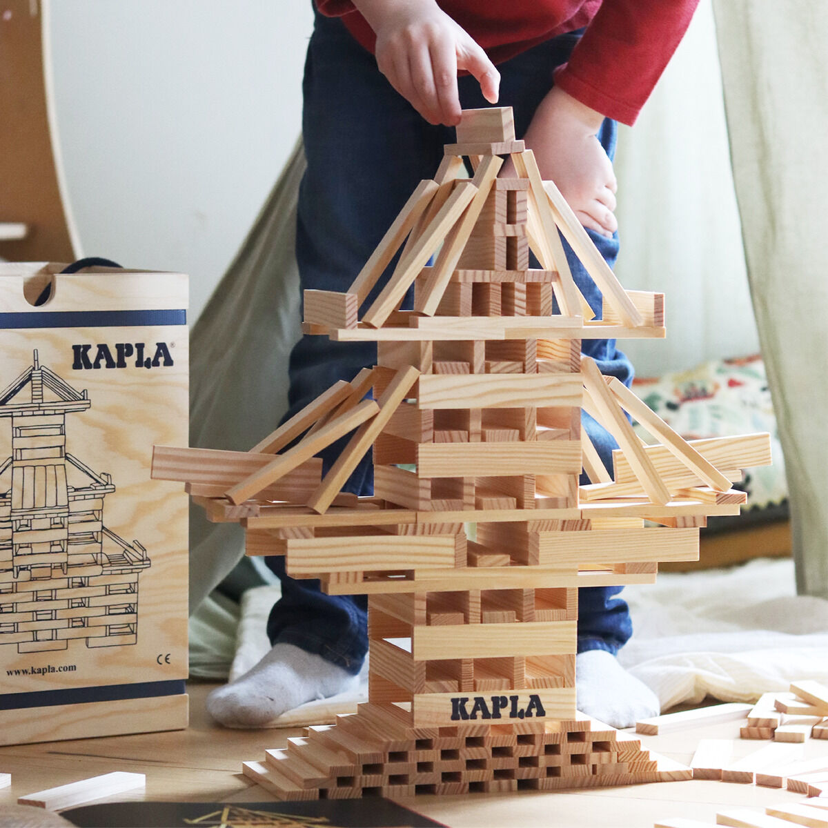 Ein Kind baut mit Holzbausteinen von Kapla ein komplexes Bauwerk. Der Hintergrund zeigt ein Zimmer. Ein Baukasten mit der Aufschrift 'Kapla' und eine Bauanleitung sind ebenfalls zu sehen.
