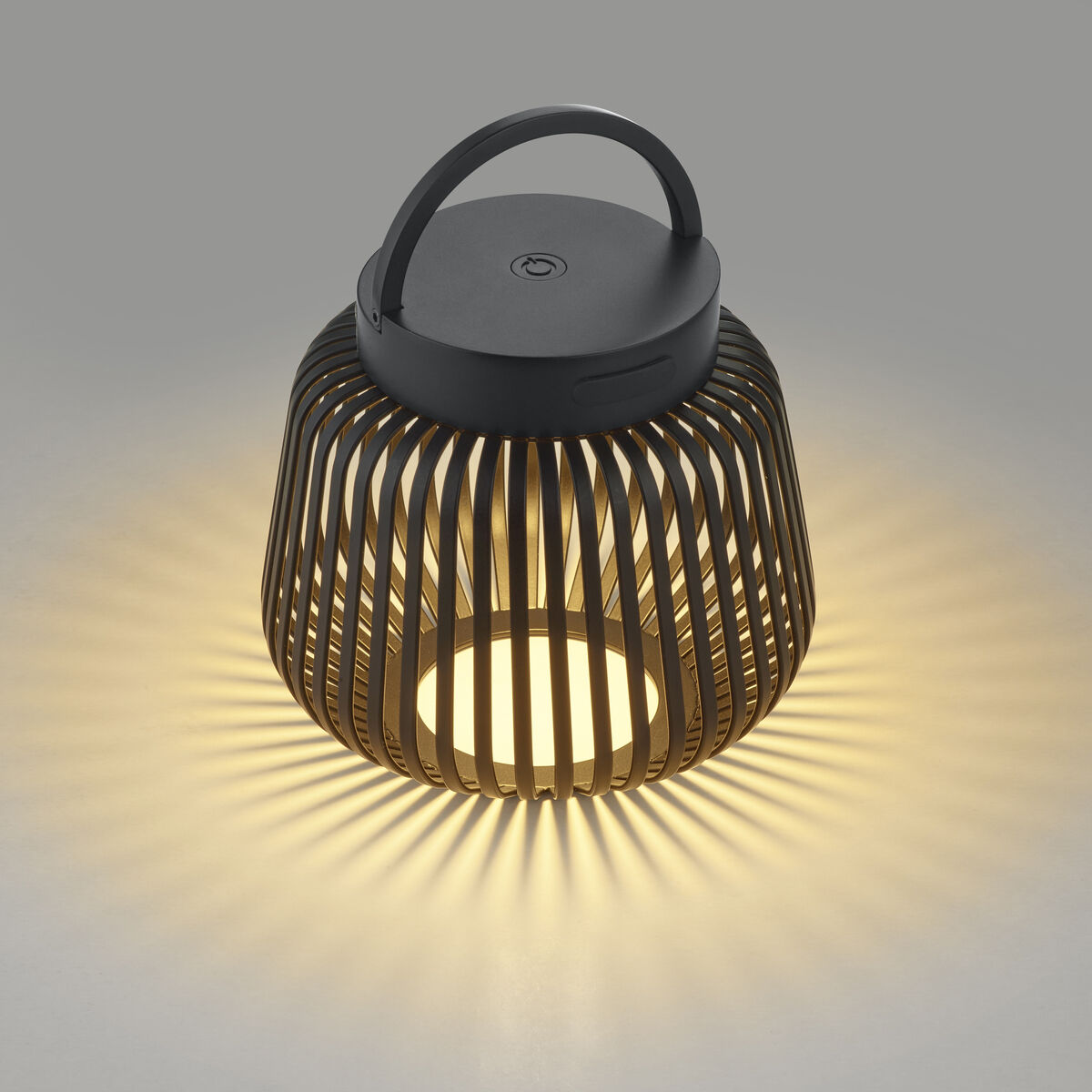 Auf dem Bild ist die LED-Leuchte "LITO" XS in Schwarz der Marke blomus zu sehen. Die Leuchte hat einen runden, offenen Metallkörper mit vertikalen Streben. Durch das warmweiße Licht entstehen strahlenförmige Muster auf der Oberfläche. Oben befindet sich ein Griff.