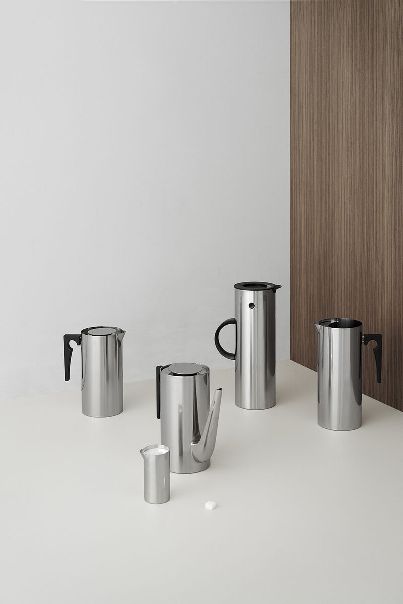 Ein Set von Arne Jacobsen Design-Kaffeekannen aus Edelstahl, minimalistisch und modern gestaltet, auf einem weißen Tisch vor einer weißen und hölzernen Wand.