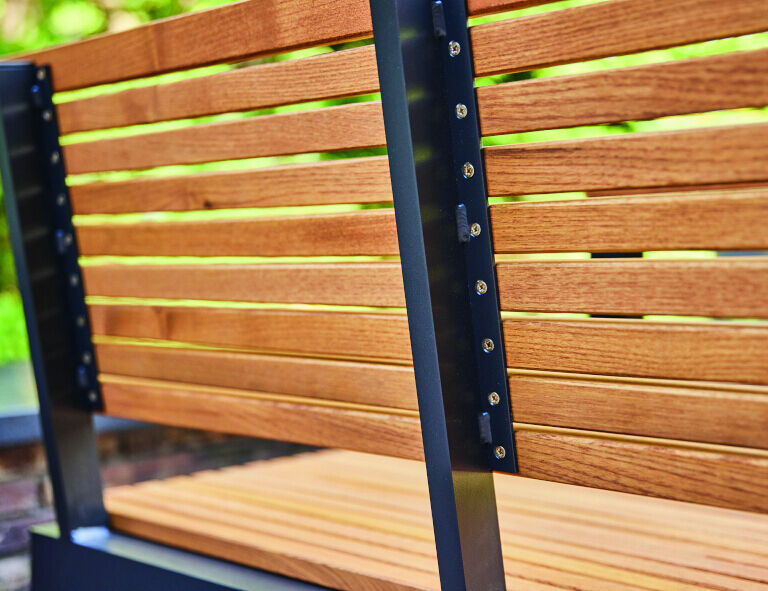 Eine moderne 2er-Bank aus Holzlatten mit schwarzem Metallrahmen steht auf einer Terrasse mit grünem Hintergrund.