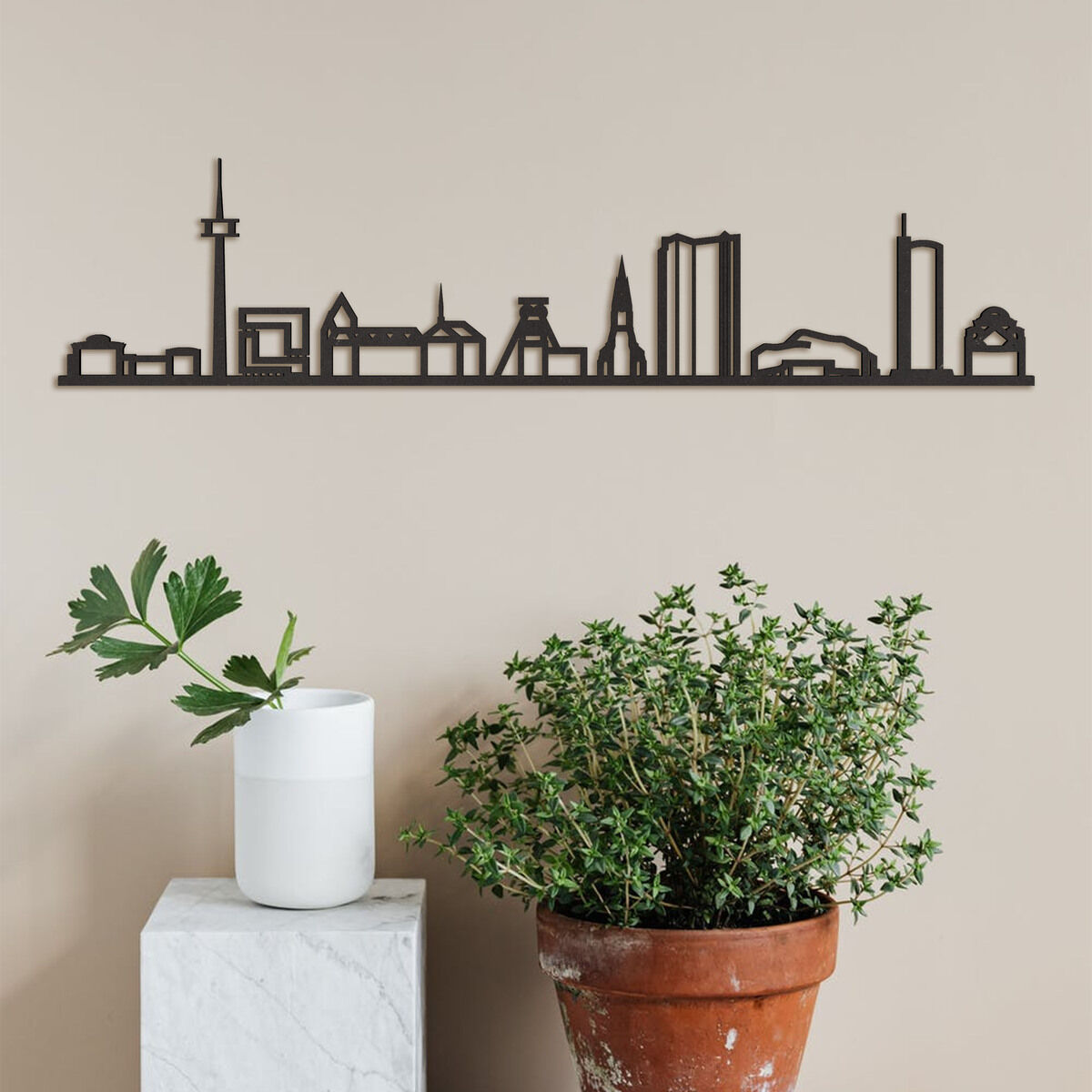 Skyline von Essen als Wanddekoration aus Holz. Die Silhouette zeigt bekannte Gebäude und Wahrzeichen der Stadt, montiert an einer beige gestrichenen Wand. Darunter sind eine verzierte Tonpflanze und eine weiße Vase mit Blättern platziert.