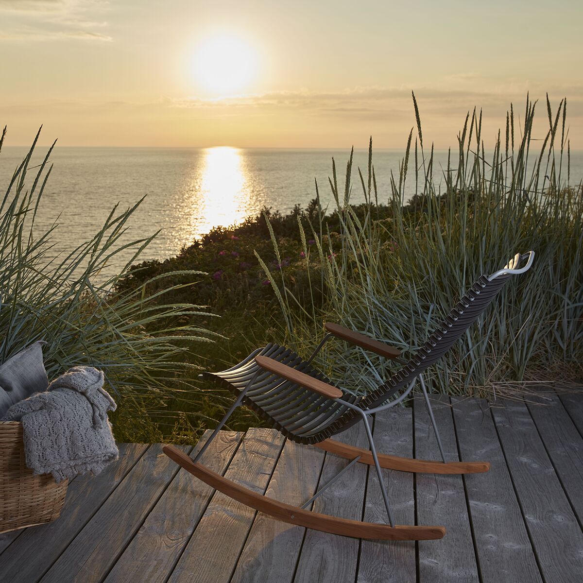 Moderner, dunkelgrauer Schaukelstuhl mit geschwungenen Holzfüßen steht auf einer Holzterrasse mit Blick auf das Meer, umgeben von hohem Gras, während im Hintergrund die Sonne untergeht. Ein Korb mit Decke und Kissen befindet sich daneben. Die Szene vermittelt Entspannung und Naturverbundenheit.