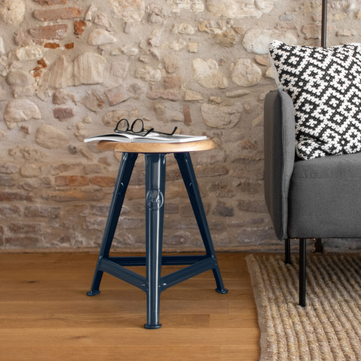 Ein stilvoller blauer Rowac-Schemel im Industrial-Design steht neben einem grauen Sofa und einem beigen Teppich. Auf dem Schemel liegt eine Brille und eine Zeitschrift, während ein gemustertes Kissen das Sofa ziert. Die Wand im Hintergrund besteht aus rustikalen Steinen.