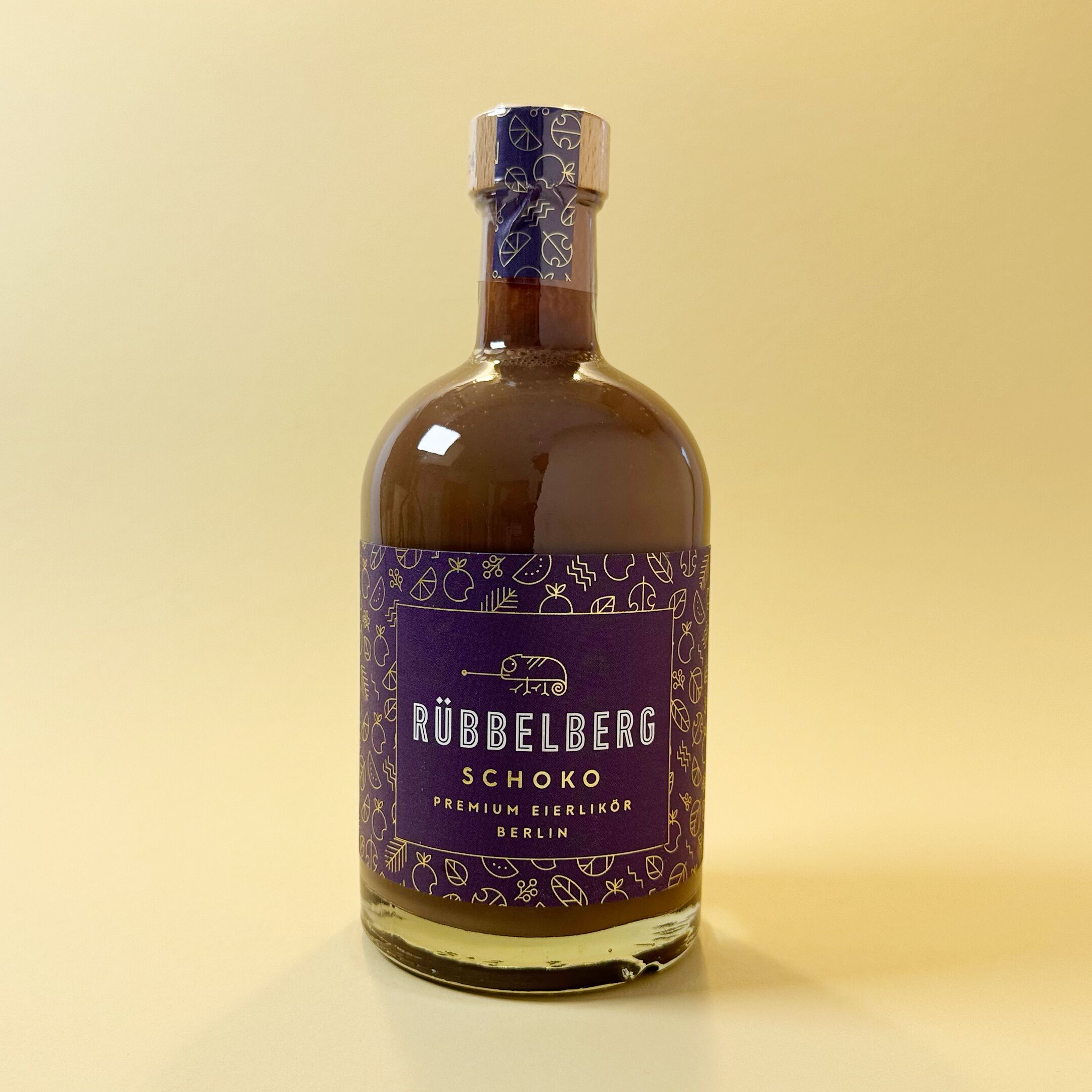 Eine Flasche Rübelberg Premium Eierlikör Schoko steht vor einem neutralen Hintergrund. Die Flasche ist mit einem lila Etikett mit goldenen und weißen grafischen Elementen gestaltet. Die bernsteinfarbene Flüssigkeit ist durch das Glas deutlich sichtbar. Der Eierlikör ist speziell für Genießer von Schokoladenlikör und hochwertige Spirituosen.