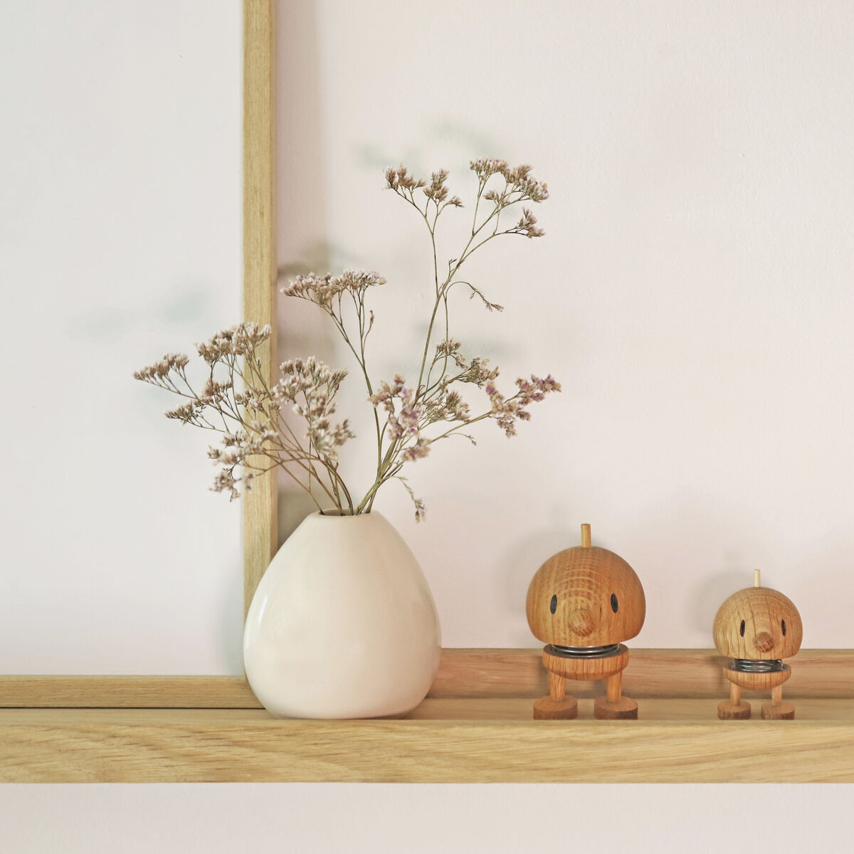 Zwei Hoptimist-Figuren aus Eichenholz der ZEIT Sonderedition stehen nebeneinander auf einem hellen Holzregal. Links daneben befindet sich eine weiße Vase mit zarten Trockenblumen, rechts ein eleganter Kerzenständer mit weißer Kerze. Das Arrangement ist in hellen, natürlichen Farben gehalten und vermittelt ein minimalistisches, skandinavisches Wohngefühl.