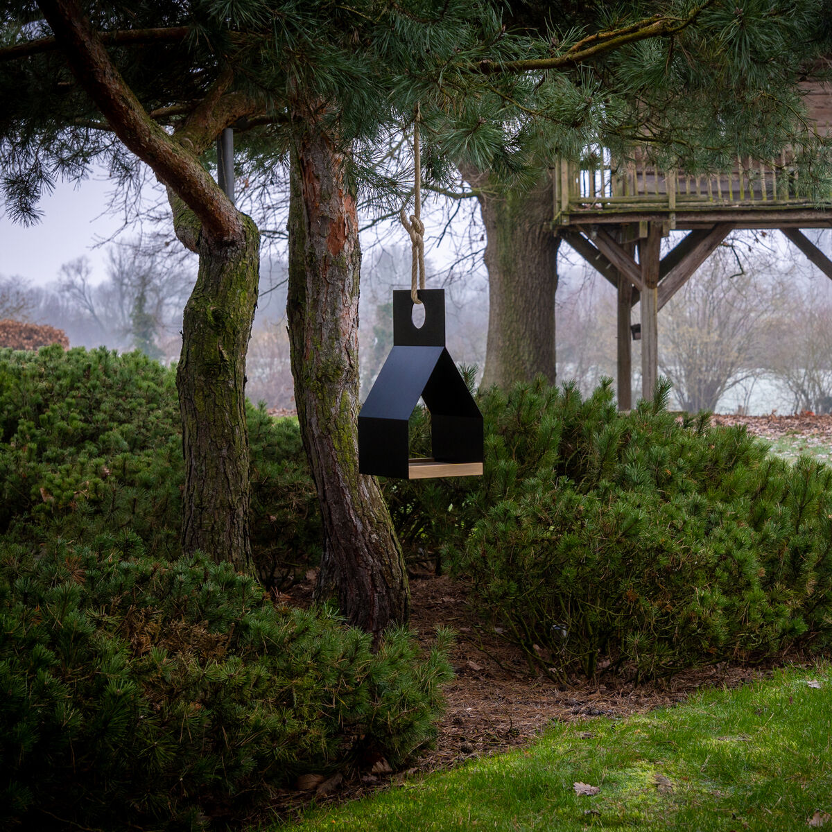 Im Vordergrund hängt das moderne Vogelhaus »FLY« von finbeaux am Baum zwischen dichten immergrünen Büschen. Im Hintergrund steht ein hölzernes Baumhaus mit Leiter. Die Szene zeigt einen gepflegten Garten an einem nebligen Tag.