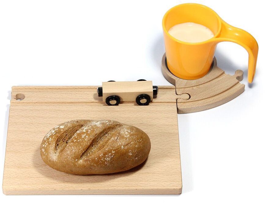 Ein Frühstücksbrett aus Holz in der Form einer Eisenbahn, darauf liegt ein Laib Brot. Daneben steht eine gelbe Tasse mit Milch auf einem Holzteilstück, das wie eine Gleisschiene geformt ist. Ein kleines Holzeisenbahn-Spielzeug befindet sich auf dem Brett.