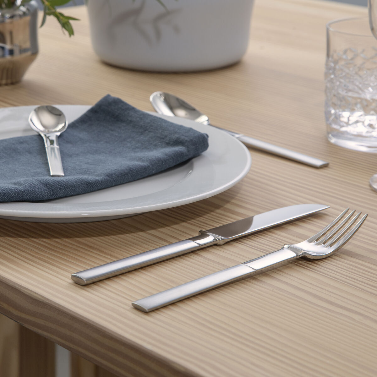 Ein moderner gedeckter Tisch mit einem Besteckset aus Edelstahl, das neben einem weißen Teller mit einer blauen Serviette arrangiert ist. Im Hintergrund sind dekorative Elemente wie Vasen und ein Glas zu sehen.