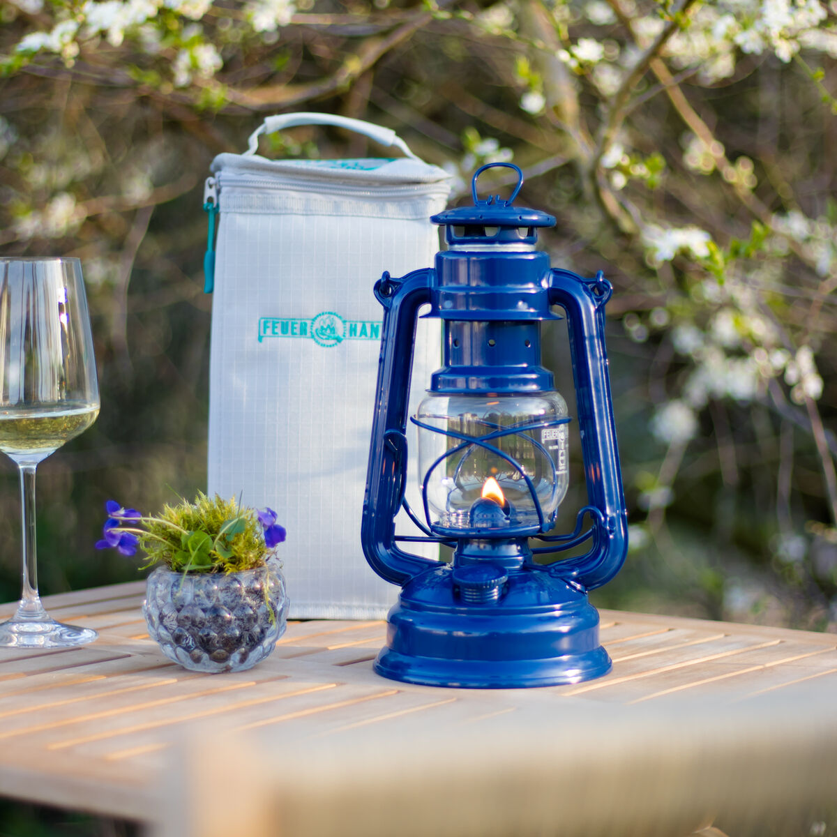 Eine blaue Sturmlaterne aus Stahl steht auf einem Holztisch im Freien neben einem Glas Weißwein und einem kleinen Blumentopf. Im Hintergrund sind blühende Bäume und ein weißer isolierter Getränkebehälter von Feuerhand zu sehen.
