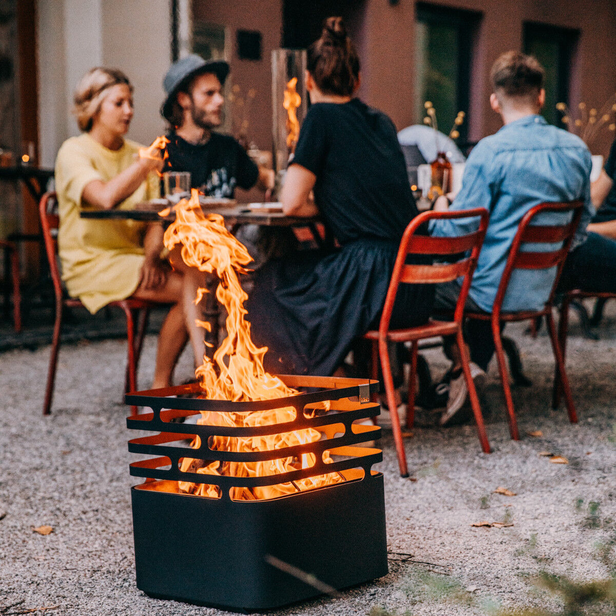 Eine Gruppe von Menschen sitzt an einem Tisch im Freien und genießt ein Essen, während eine moderne Feuerschale, »CUBE« Plancha, leuchtende Flammen in die Luft wirft.
