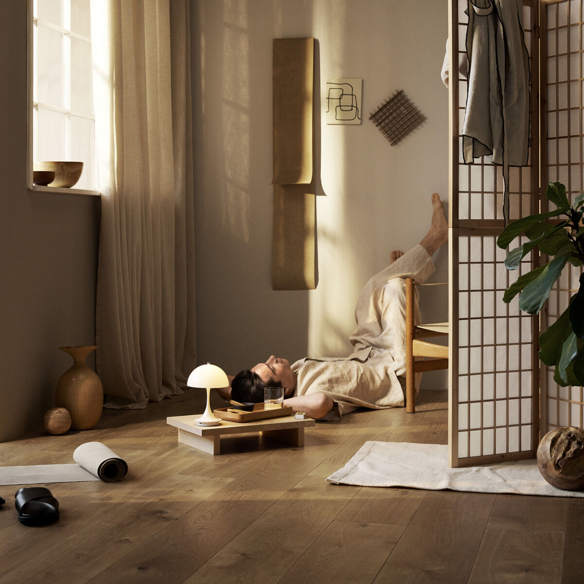 Skandinavisch eingerichtetes Wohnzimmer mit einer Panthella 160 Portable Leuchte auf einem Tisch. Der Raum ist mit Holzboden, einem Raumteiler und Pflanzen dekoriert. Weiches natürliches Licht strömt durch ein großes Fenster.