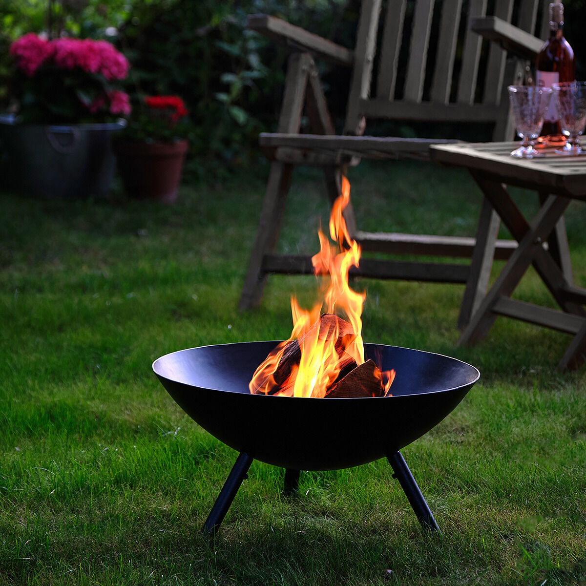 Auf einem grünen Rasen steht eine schlichte, schwarze Feuerschale »Flames« von Philippi mit loderndem Feuer, dahinter ein Holztisch mit Gläsern und Wein sowie eine Gartenbank. Im Hintergrund blühende Blumen und Pflanzen, sommerliche Atmosphäre, Dämmerung.