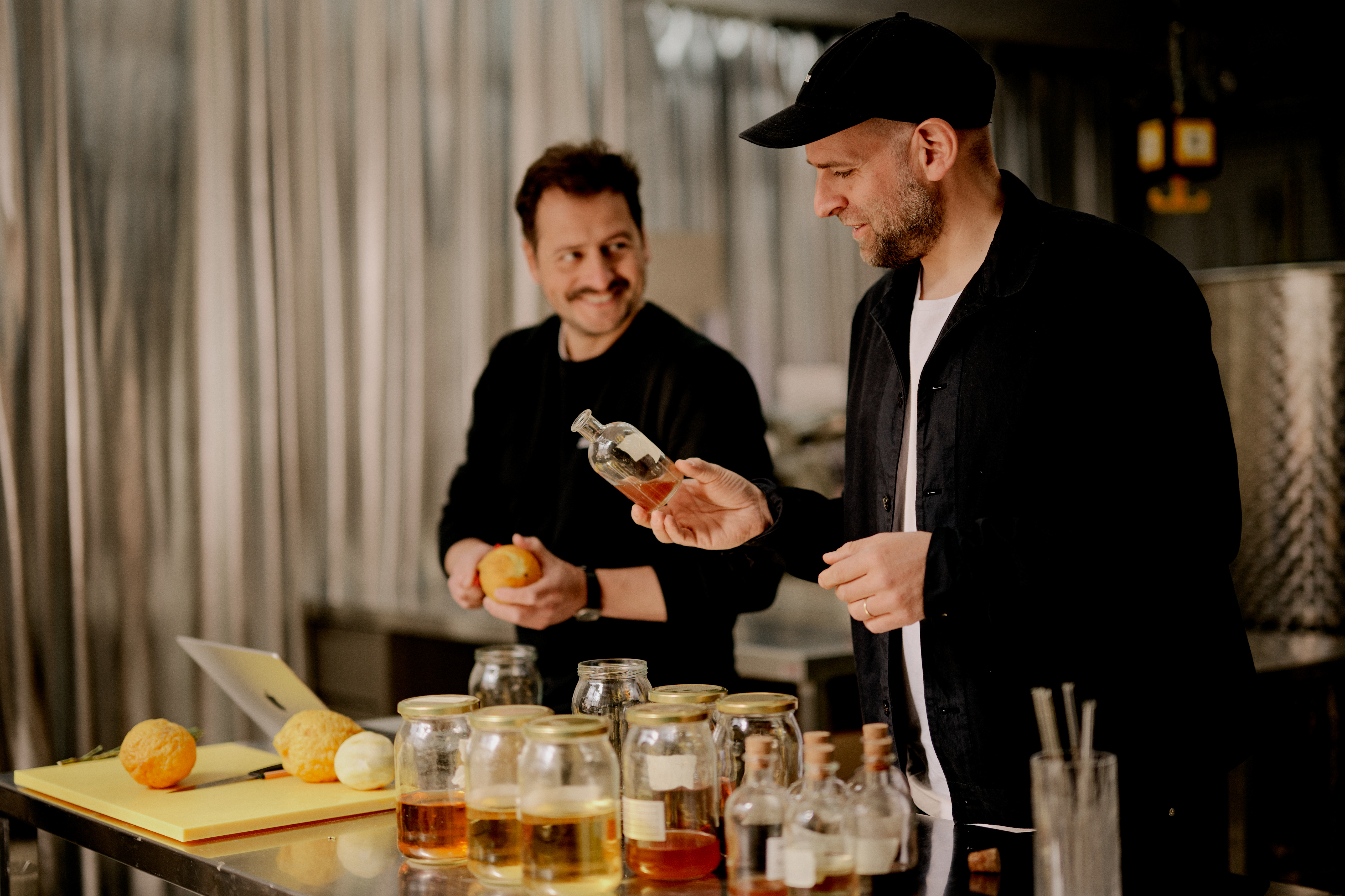 Zwei Männer in einer Küche, einer hält eine Flasche mit Flüssigkeit, der andere eine Orange, mehrere Gläser mit eingelegtem Obst auf dem Tisch