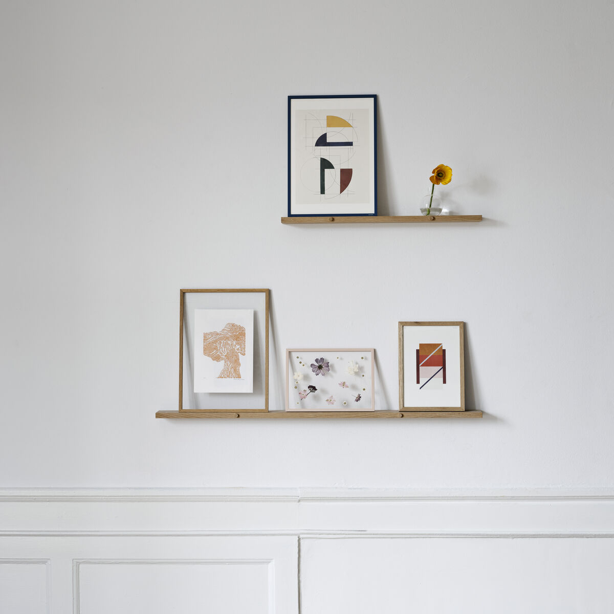 Ein minimalistisches Wandregal aus Eichenholz, das zwei dünne Regalbretter umfasst. Darauf befinden sich mehrere Bilderrahmen mit abstrakter Kunst und eine kleine Sonnenblume in einer Vase. Die Wand ist weiß gestrichen und der Boden besteht aus braunem Parkett.