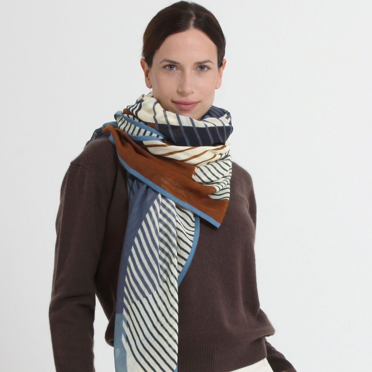 Frau trägt einen großzügigen, gestreiften Wollschal »Alma« in Blau-Braun von Shalls Barcelona. Der Schal ist 180x100 cm groß, aus 100% Schurwolle, farblich dezent mit Streifen in Blau, Weiß und Braun gehalten und locker um Hals und Schultern gelegt.