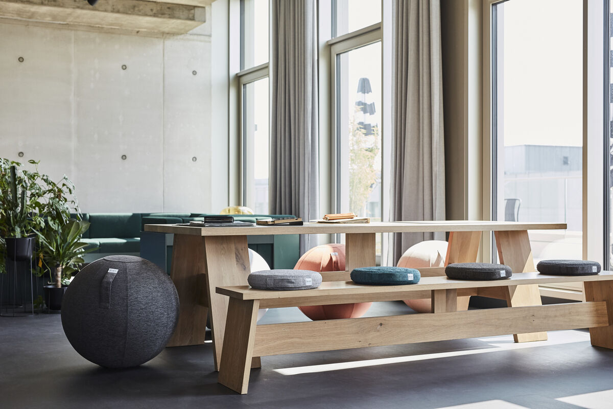 Ein modernes Wohnzimmer mit großen Fenstern, einem Holztisch und -bänken sowie mehreren VLUV-Balancekissen in Grautönen.