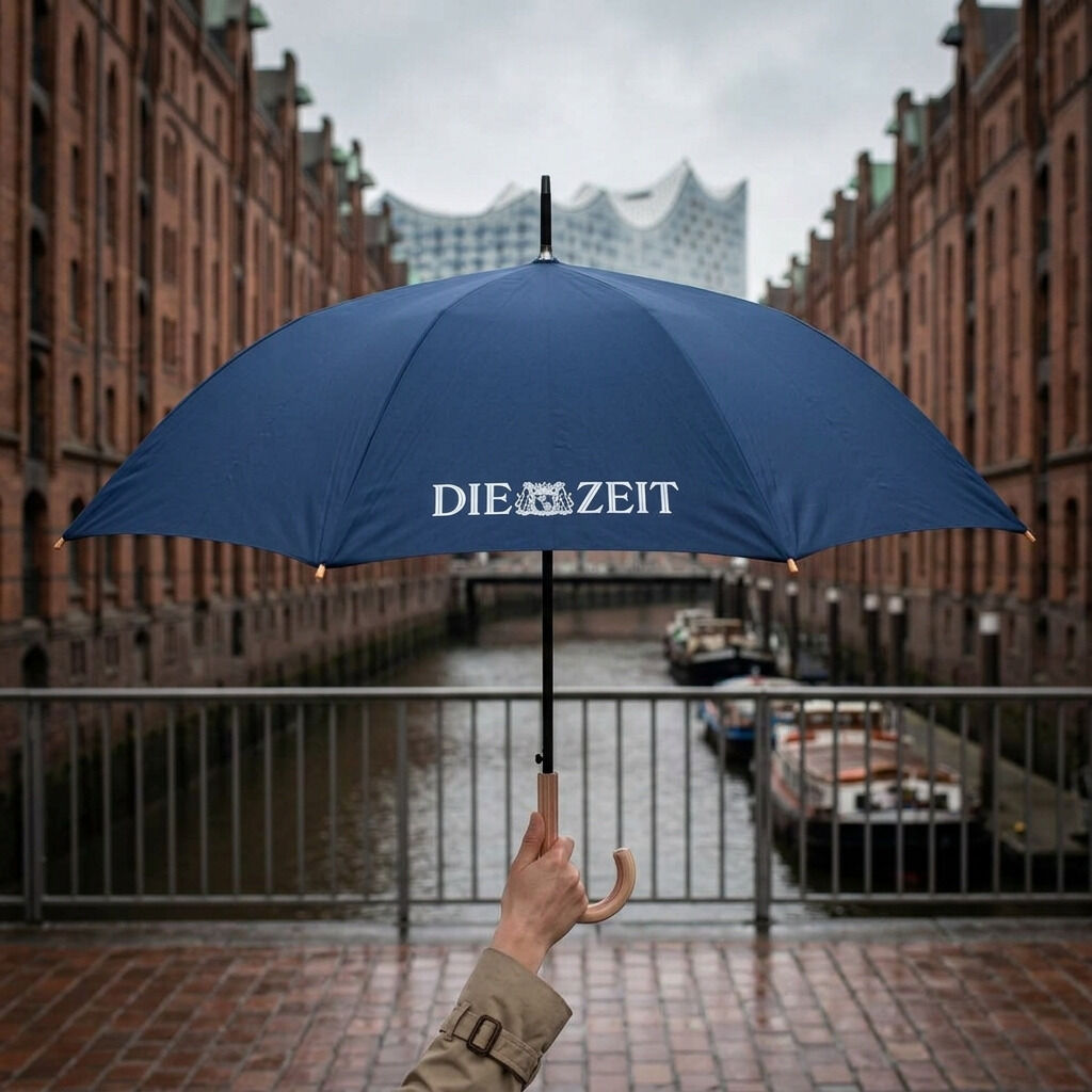 Eine Person hält einen dunkelblauen Regenschirm mit weißem DIE ZEIT-Logo und elegantem Echtholzgriff vor der historischen Kulisse der Hamburger Speicherstadt. Hinten sind Backsteingebäude, Wasser und Boote zu sehen, im Hintergrund die Elbphilharmonie.