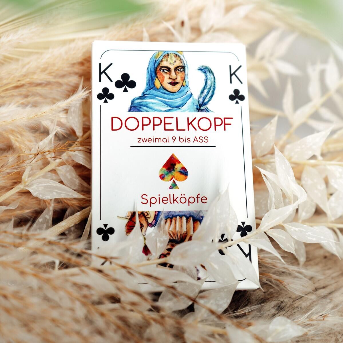 Eine Doppelkopf-Spielkarte liegt auf einem Bett aus trockenem Gras. Die Karte ist mit dem Symbol Pik und einem König dargestellt. Oben in der Mitte steht 'Doppelkopf zweimal 9 bis Ass' und es gibt ein farbenreiches Logo von 'Spielköpfe'.