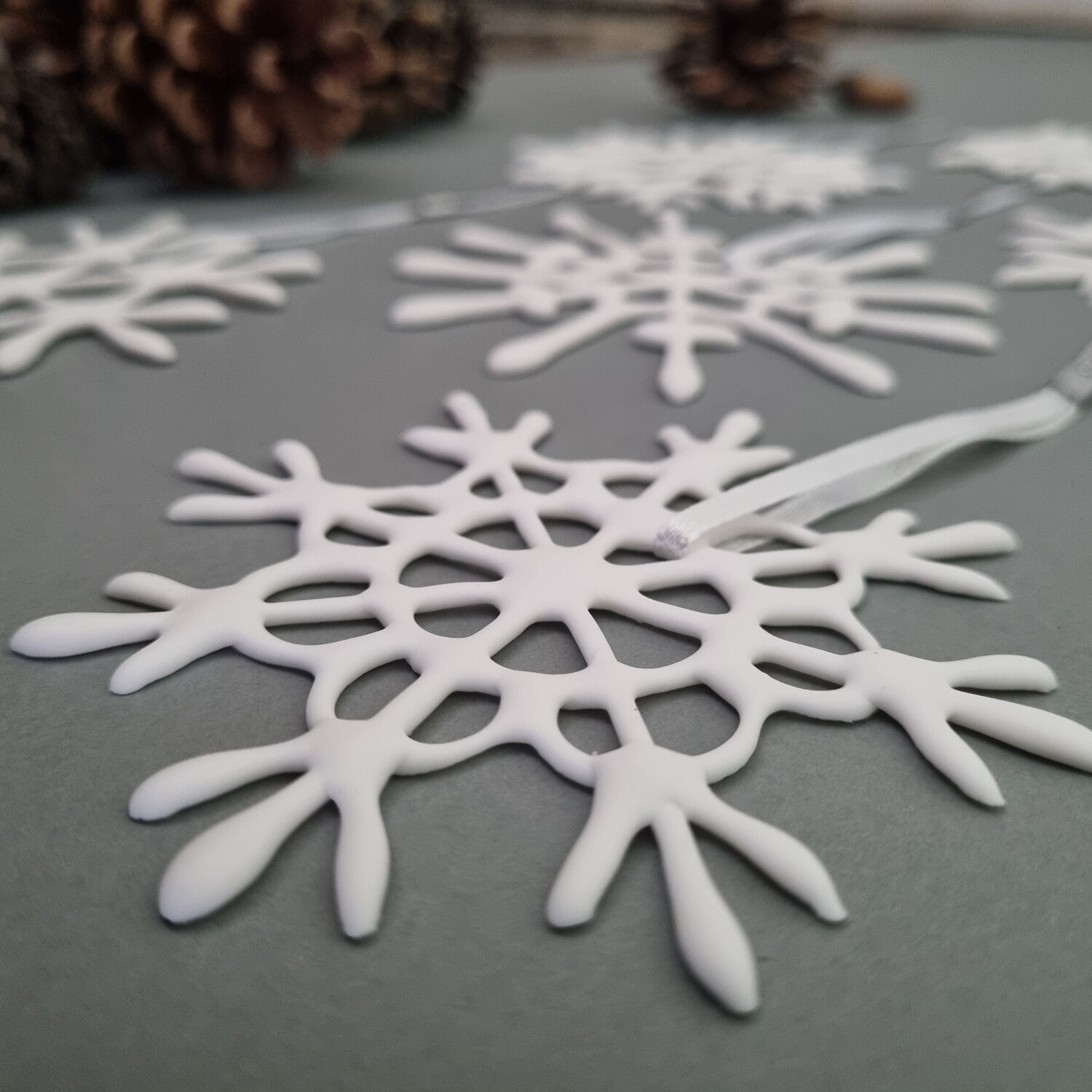 Sechs weiße Schneekristall-Anhänger aus Kunststoff liegen dekorativ auf einer grauen Oberfläche. Im Hintergrund sind einige Zapfen zu sehen. Die Schneeflocken sind filigran gearbeitet und mit weißen Bändern versehen, ideal für festliche Dekoration in der Winter- und Weihnachtszeit.