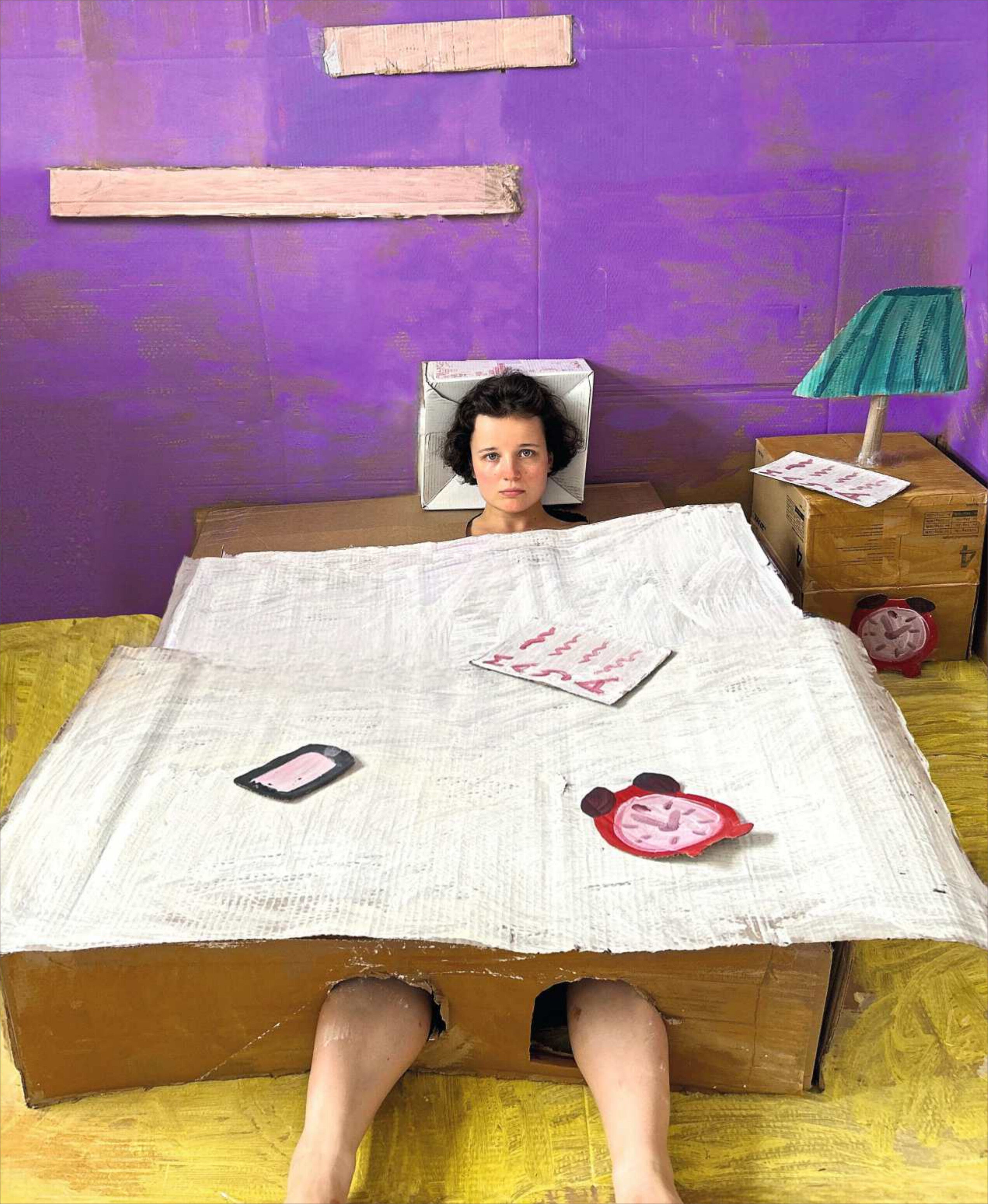 Eine junge Person liegt in einem Bett aus Kartons in einem Zimmer mit lilafarbenen Wänden und gelbem Boden. Neben dem Bett befinden sich ein Nachttisch, darauf eine grüne Lampe sowie weitere Kartonmöbel. Auf Bettdecke und Möbeln liegen Zeitschriften und eine Uhr. Die Szenerie vermittelt das Thema Wohnen auf kreative, provisorische Weise.