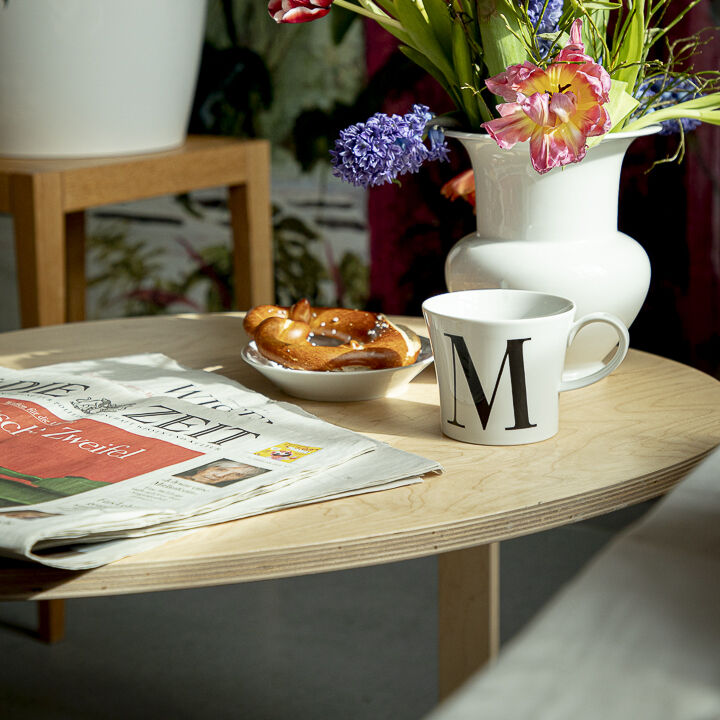 Auf einem runden Holztisch stehen eine weiße Frühstückstasse mit einem schwarzen Buchstaben 'M', eine Tageszeitung, ein Brezel und ein weißer Krug mit einem bunten Blumenstrauß. Im Hintergrund sind grüne Pflanzen sichtbar.