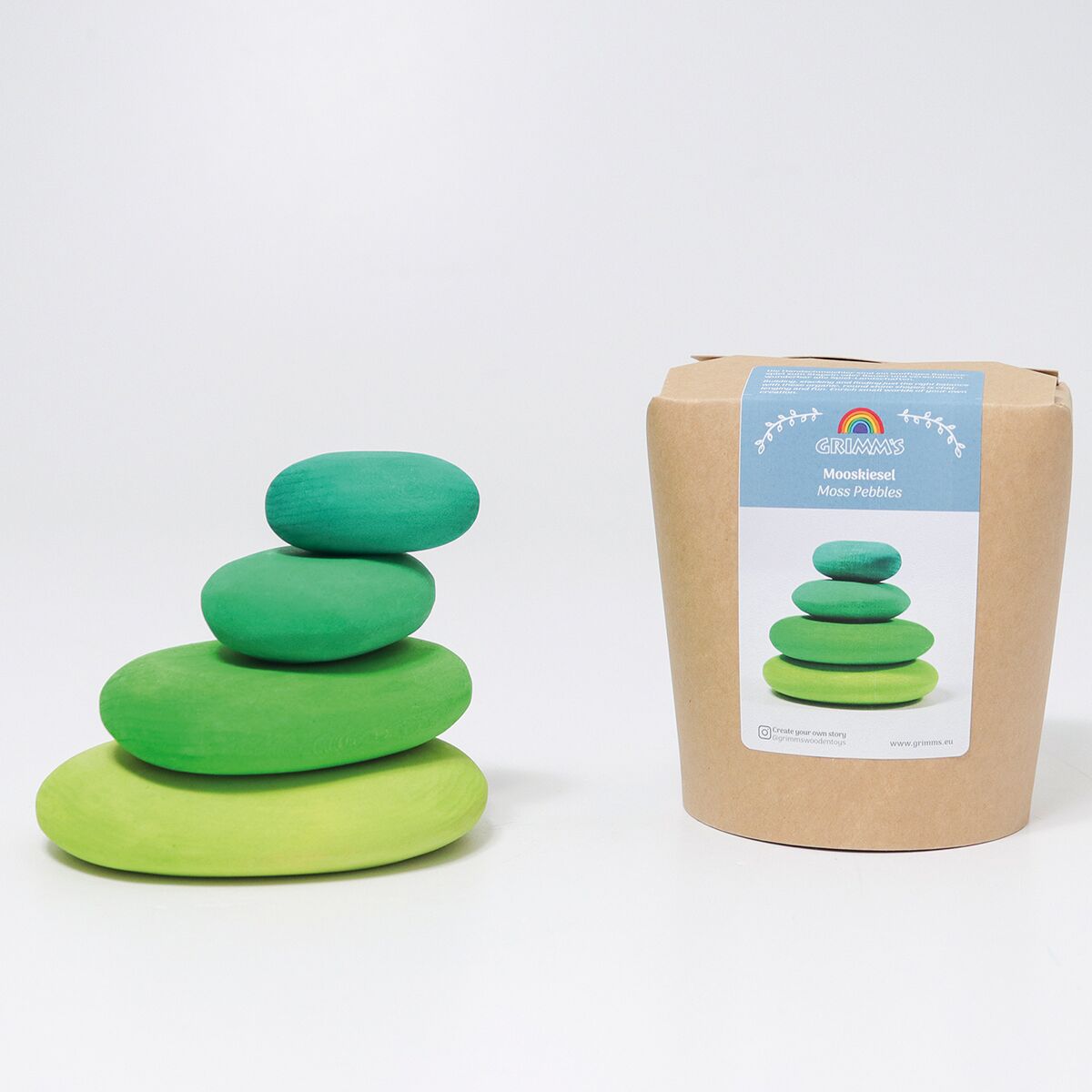 Vier unterschiedlich große, grüne, tropfenförmige Holzsteine liegen zu einem kleinen Turm gestapelt neben einer naturfarbenen Verpackung. Die Verpackung zeigt eine Abbildung der Holzsteine und trägt die Marke GRIMM’S sowie den Produktnamen Mooskiesel.