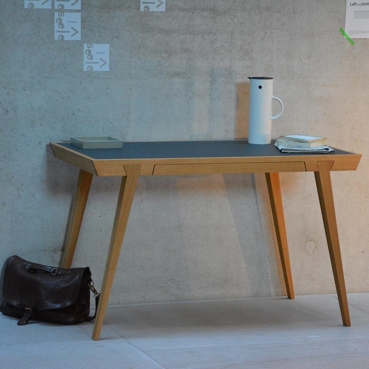 Schreibtisch »Desk«, ein moderner, minimalistisch gestalteter Bürotisch aus Holz mit einer grauen Arbeitsfläche. Auf dem Tisch stehen eine weiße Kanne und Bücher. Eine braune Ledertasche liegt auf dem Boden daneben. Der Hintergrund ist eine graue Wand.