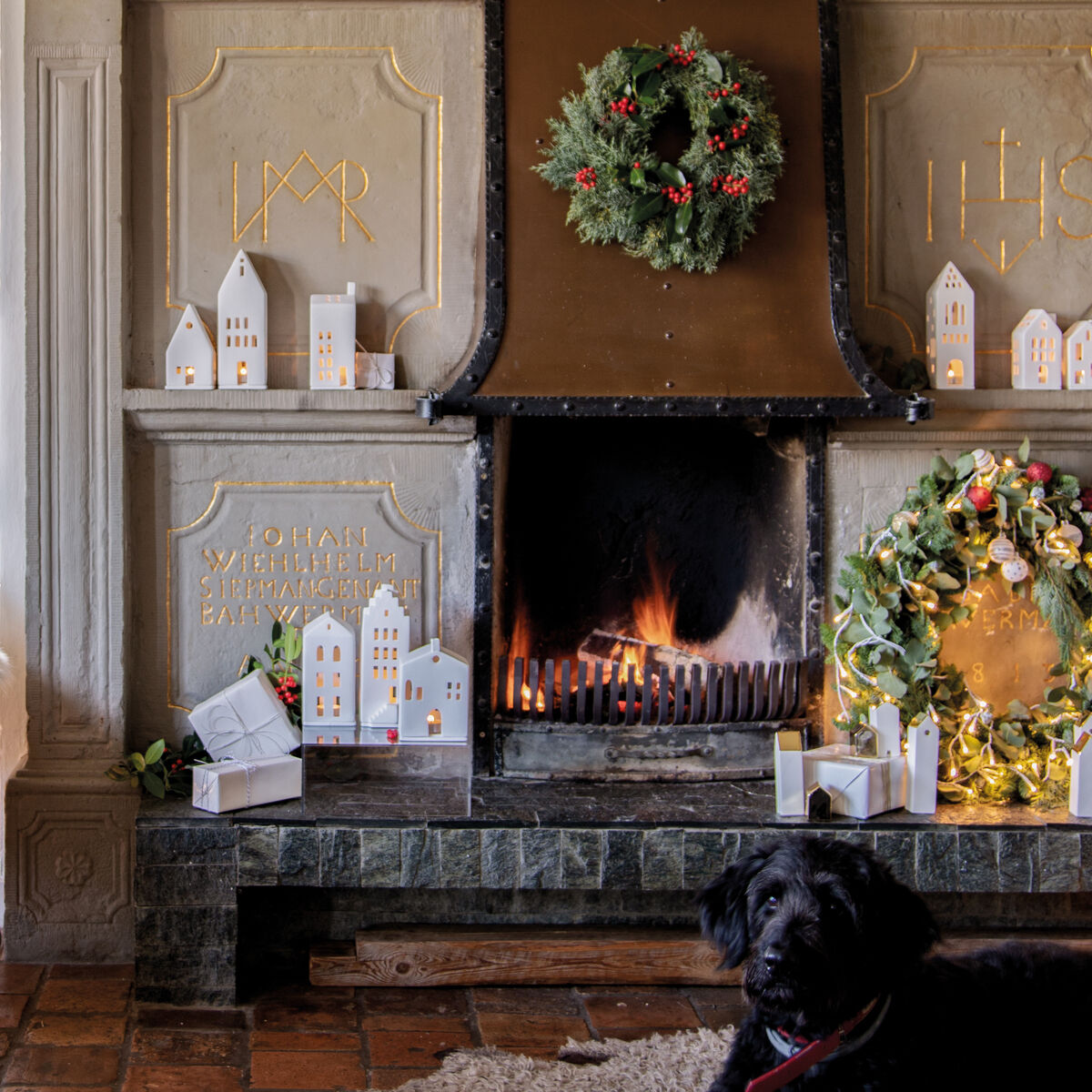 Gemütlicher Innenraum mit einem brennenden Kamin, umgeben von festlichen Dekorationen wie weißem Lichthaus im Giebeldach-Stil, grünen Kränzen und Kerzen. Ein schwarzer Hund liegt auf einem Teppich vor dem Kamin.