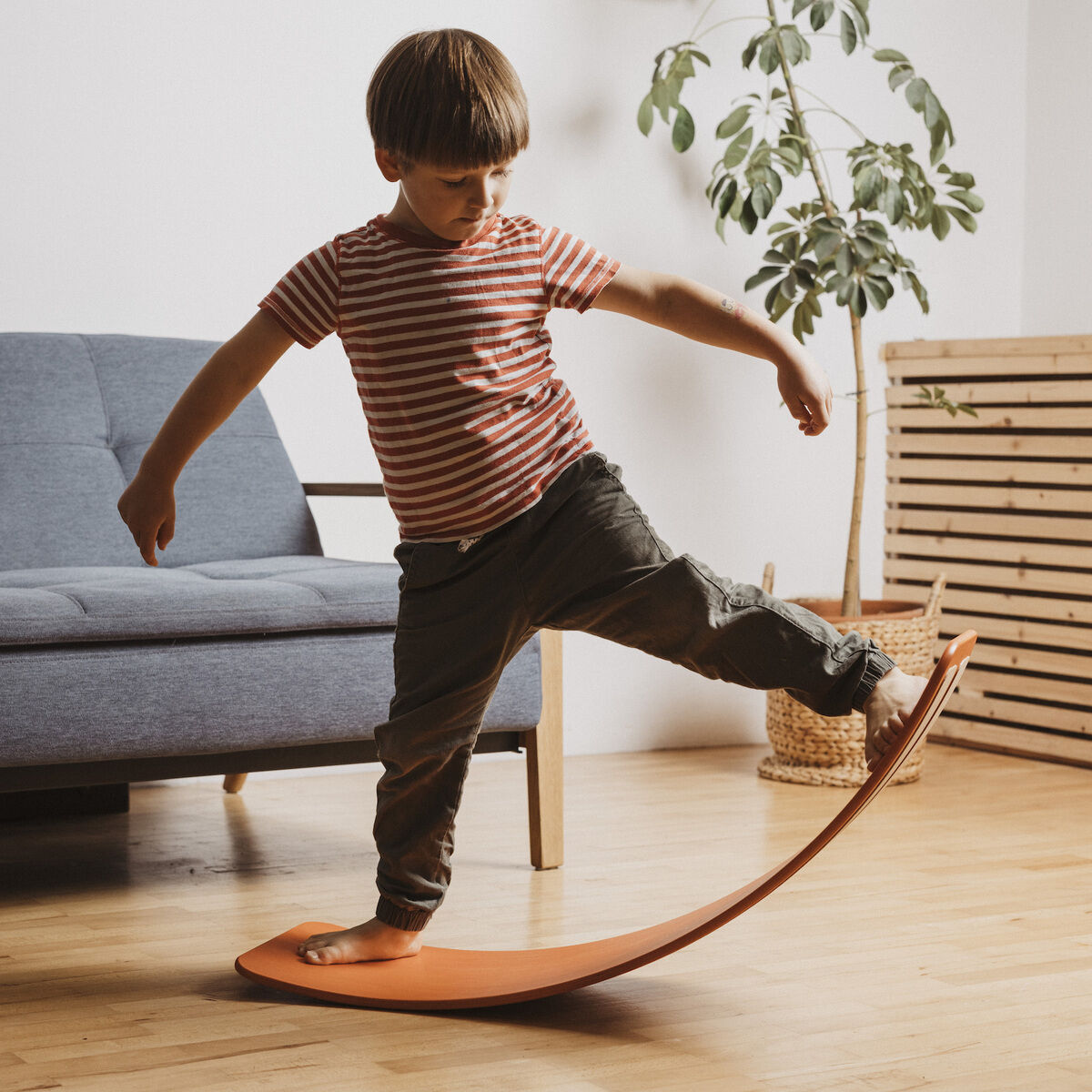 Ein Kind balanciert auf einem roten Balanceboard im Wohnzimmer, das mit einem grauen Sofa, einem Holzboden und einer Pflanze im Hintergrund ausgestattet ist.