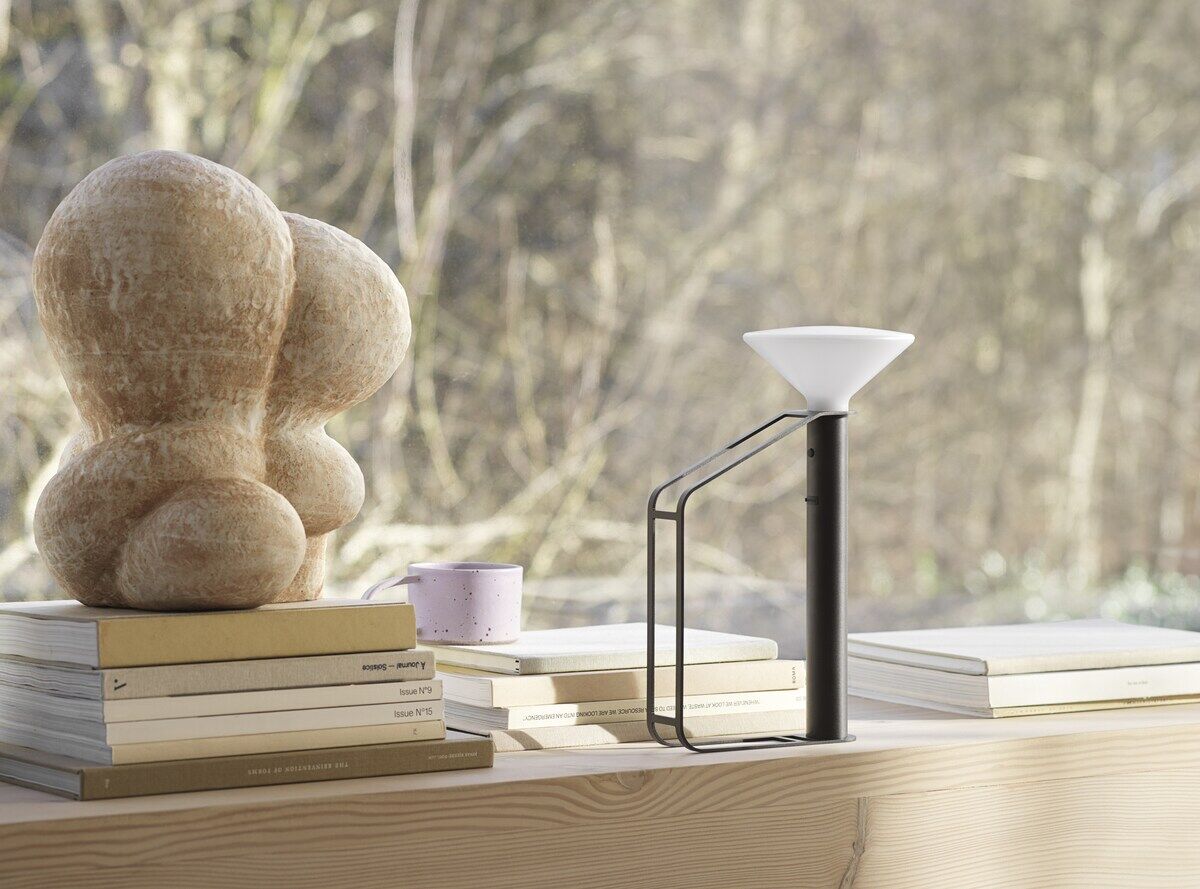 Eine tragbare Lampe namens 'Piton' steht auf einem Tisch neben einem Stapel Bücher und einer Skulptur. Die Lampe hat ein modernes und minimalistisches Design mit einem schwarzen Ständer und einem weißen Lampenschirm. Im Hintergrund sind Bäume zu sehen, die durch ein großes Fenster hindurch sichtbar sind.