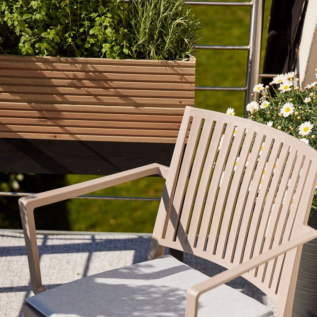 Ein Hochbeet mit Kräutern und Blumen steht auf einem Balkon oder einer Terrasse. Es gibt einen beige Gartenstuhl und einen Mann, der ein Buch liest. Der Hintergrund ist grün und zeigt einen sonnigen Tag.