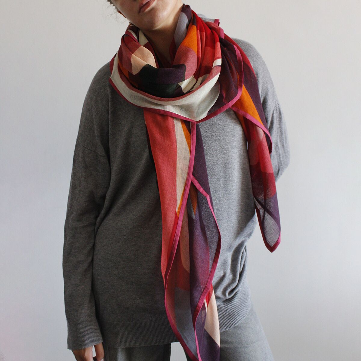 Ein modischer Wollschal namens »Yara« in verschiedenen Rottönen mit abstraktem Muster und transparentem Look liegt locker um den Hals einer Person, die einen grauen Pullover trägt. Das Accessoire hebt sich durch seine lebhaften Farben hervor und wirkt weich sowie elegant.